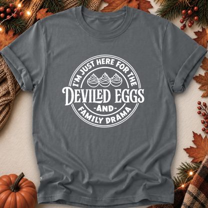 A dark gray crewneck t-shirt with a white circular graphic that reads 'I'M JUST HERE FOR THE DEVILED EGGS AND FAMILY DRAMA' with three egg illustrations, displayed in a flat-lay autumn setting with pumpkins, pine branches, and leaves.