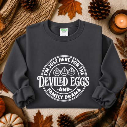 A dark charcoal gray crewneck sweatshirt with a white graphic print reading 'I'M JUST HERE FOR THE DEVILED EGGS AND FAMILY DRAMA' with three devil horns, styled in a flat lay with autumn props including pinecones, a candle, and a knit blanket.
