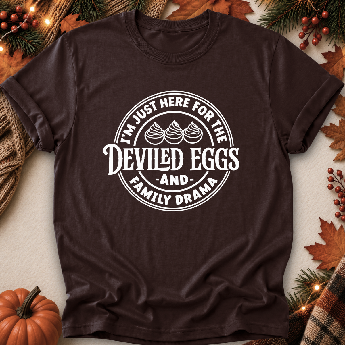 A dark brown short-sleeve t-shirt with a white circular graphic that reads 'I'M JUST HERE FOR THE DEVILED EGGS AND FAMILY DRAMA' with three illustrated deviled eggs. The shirt is laid flat on a light beige surface surrounded by autumn-themed props including a small pumpkin, pine branches, leaves, and string lights.