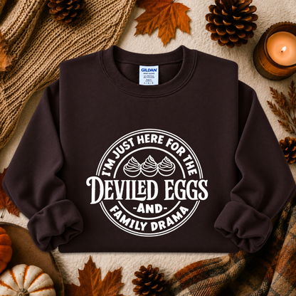 A dark brown crewneck sweatshirt with a white circular graphic reading 'I'M JUST HERE FOR THE DEVILED EGGS AND FAMILY DRAMA' laid flat on a cozy autumn-themed background with pinecones, leaves, a candle, and a knit blanket.