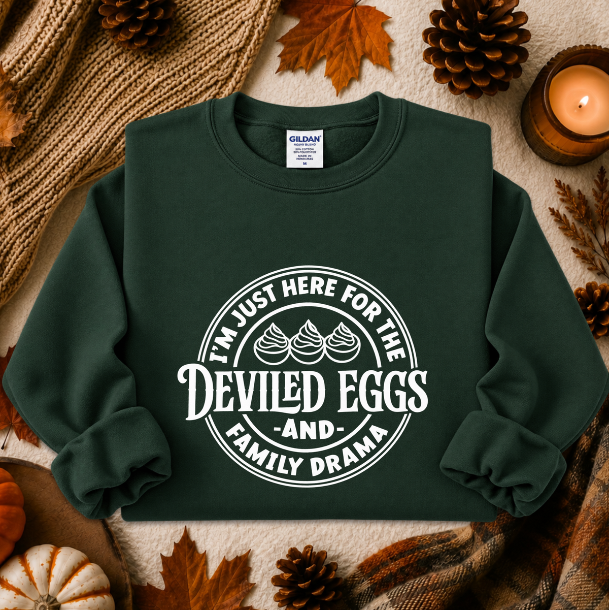 A dark green crewneck sweatshirt with a white graphic print reading 'I'M JUST HERE FOR THE DEVILED EGGS AND FAMILY DRAMA' with three devil-egg illustrations, laid flat on a cozy autumn-themed background with leaves, pinecones, a candle, and a knit blanket.