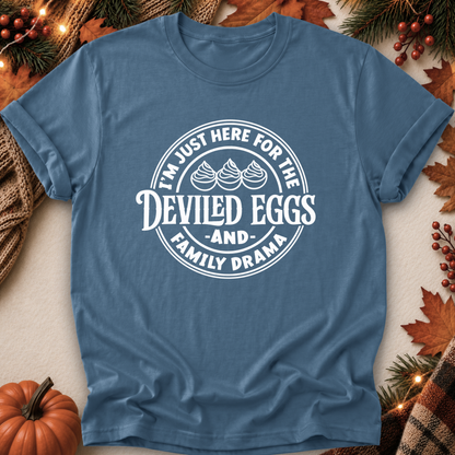 A blue short-sleeve crewneck t-shirt with a white circular graphic reading 'I'M JUST HERE FOR THE DEVILED EGGS AND FAMILY DRAMA' with three egg illustrations, laid flat on a neutral background with autumn decorations including a pumpkin, pine branches, and leaves.