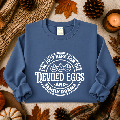A blue crewneck sweatshirt with a white graphic print reading 'I'M JUST HERE FOR THE DEVILED EGGS AND FAMILY DRAMA' laid flat on a cozy autumn-themed background with pinecones, leaves, a candle, and a knit blanket.