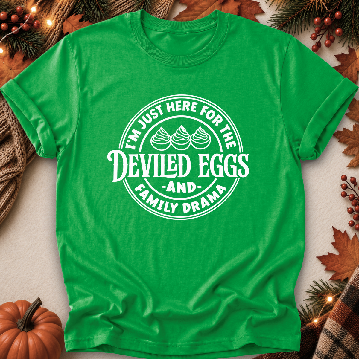 A bright green short-sleeve t-shirt with a white circular graphic reading 'I'M JUST HERE FOR THE DEVILED EGGS AND FAMILY DRAMA' with three devil-egg illustrations, laid flat on a neutral surface with autumn decorations including leaves, pumpkins, pine branches, and string lights.