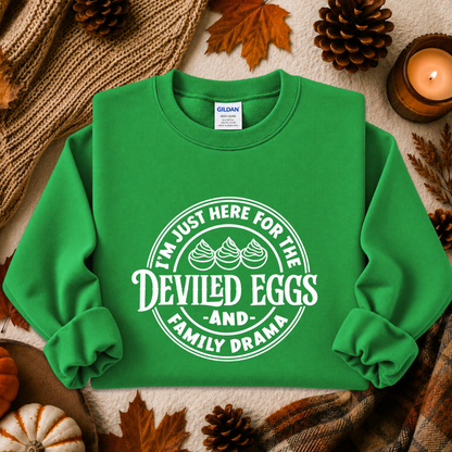 A bright green crewneck sweatshirt with a white graphic print that reads 'I'M JUST HERE FOR THE DEVILED EGGS AND FAMILY DRAMA' with three deviled eggs illustration. The sweatshirt is styled in a flat-lay autumn setting with pinecones, a candle, and a knit blanket.