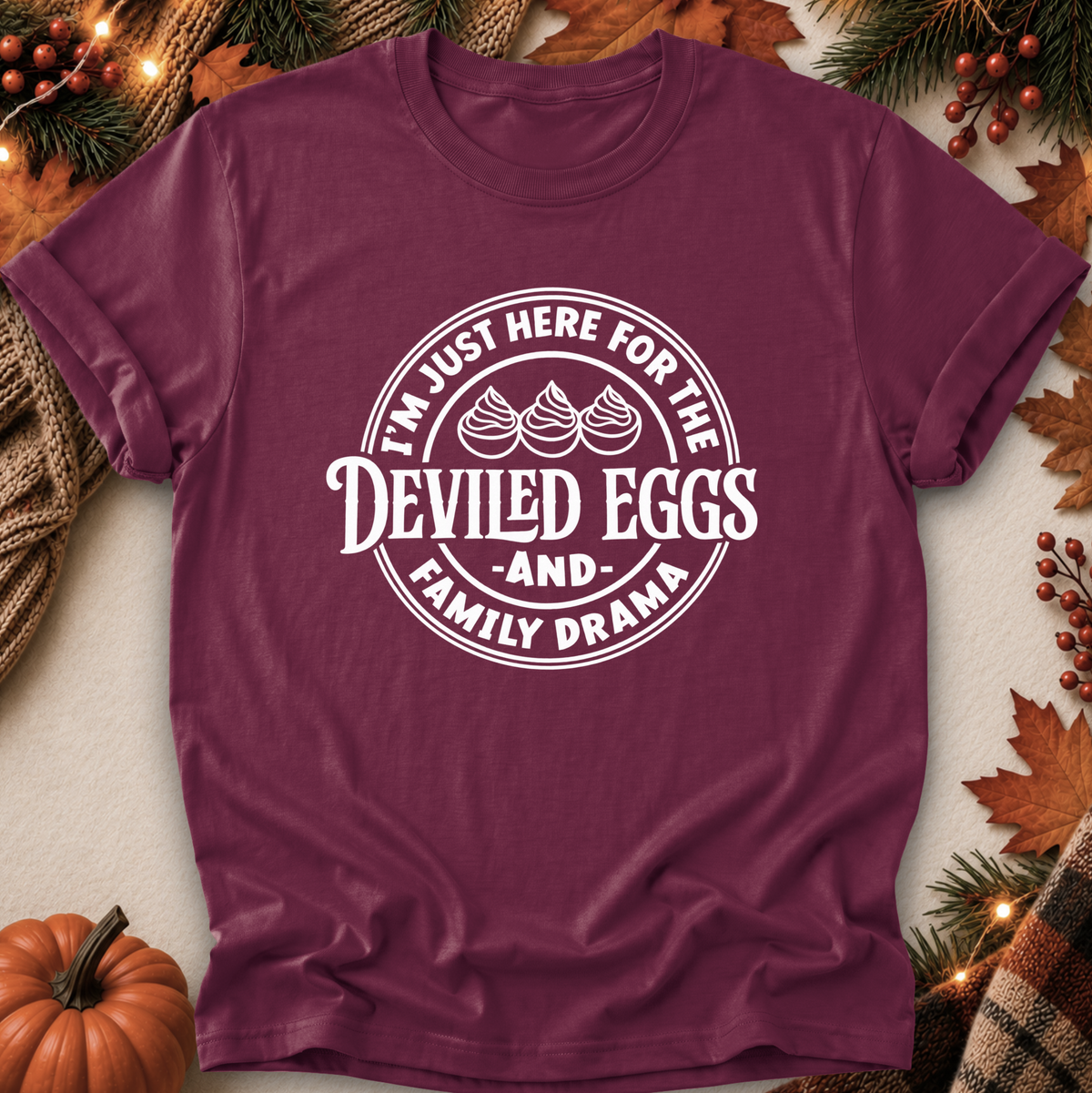 A maroon short-sleeve crewneck t-shirt with a white circular graphic reading 'I'M JUST HERE FOR THE DEVILED EGGS AND FAMILY DRAMA' with three devil-egg illustrations, displayed in a flat-lay autumn setting with pumpkins, pine branches, and fall leaves.