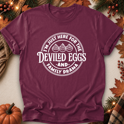 A maroon short-sleeve crewneck t-shirt with a white circular graphic reading 'I'M JUST HERE FOR THE DEVILED EGGS AND FAMILY DRAMA' with three devil-egg illustrations, displayed in a flat-lay autumn setting with pumpkins, pine branches, and fall leaves.