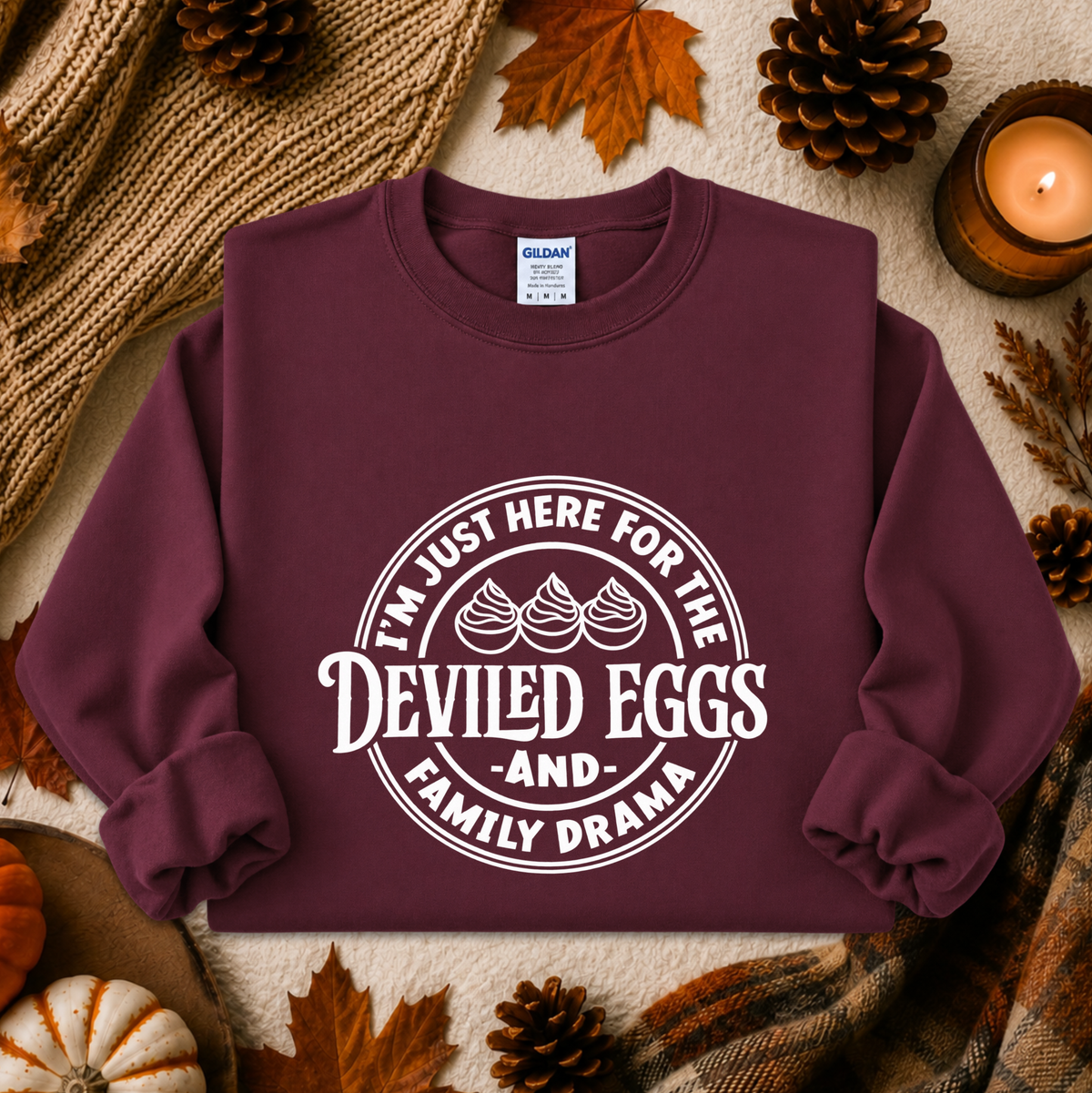 A burgundy crewneck sweatshirt with a white graphic print reading 'I'M JUST HERE FOR THE DEVILED EGGS AND FAMILY DRAMA' with three egg illustrations, styled in an autumn flat-lay with a knit blanket, pinecones, leaves, and a lit candle.
