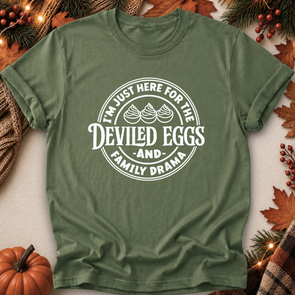 An olive green short-sleeve t-shirt with a white circular graphic that reads 'I'M JUST HERE FOR THE DEVILED EGGS AND FAMILY DRAMA' with three devil-egg illustrations, laid flat on a light background with autumn decorations like a pumpkin, pine branches, and leaves.