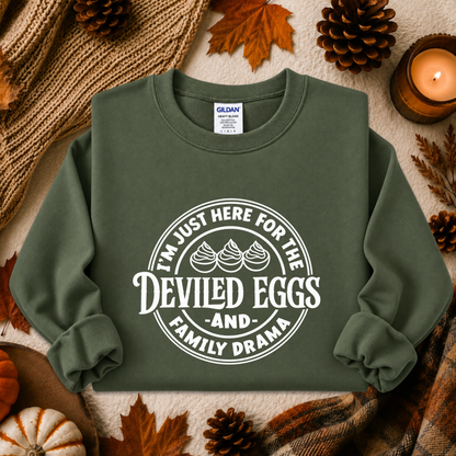 An olive green crewneck sweatshirt with a white graphic print reading 'I'M JUST HERE FOR THE DEVILED EGGS AND FAMILY DRAMA' surrounded by three devil horns, styled in an autumn flat-lay with pinecones, leaves, a candle, and a knit blanket.