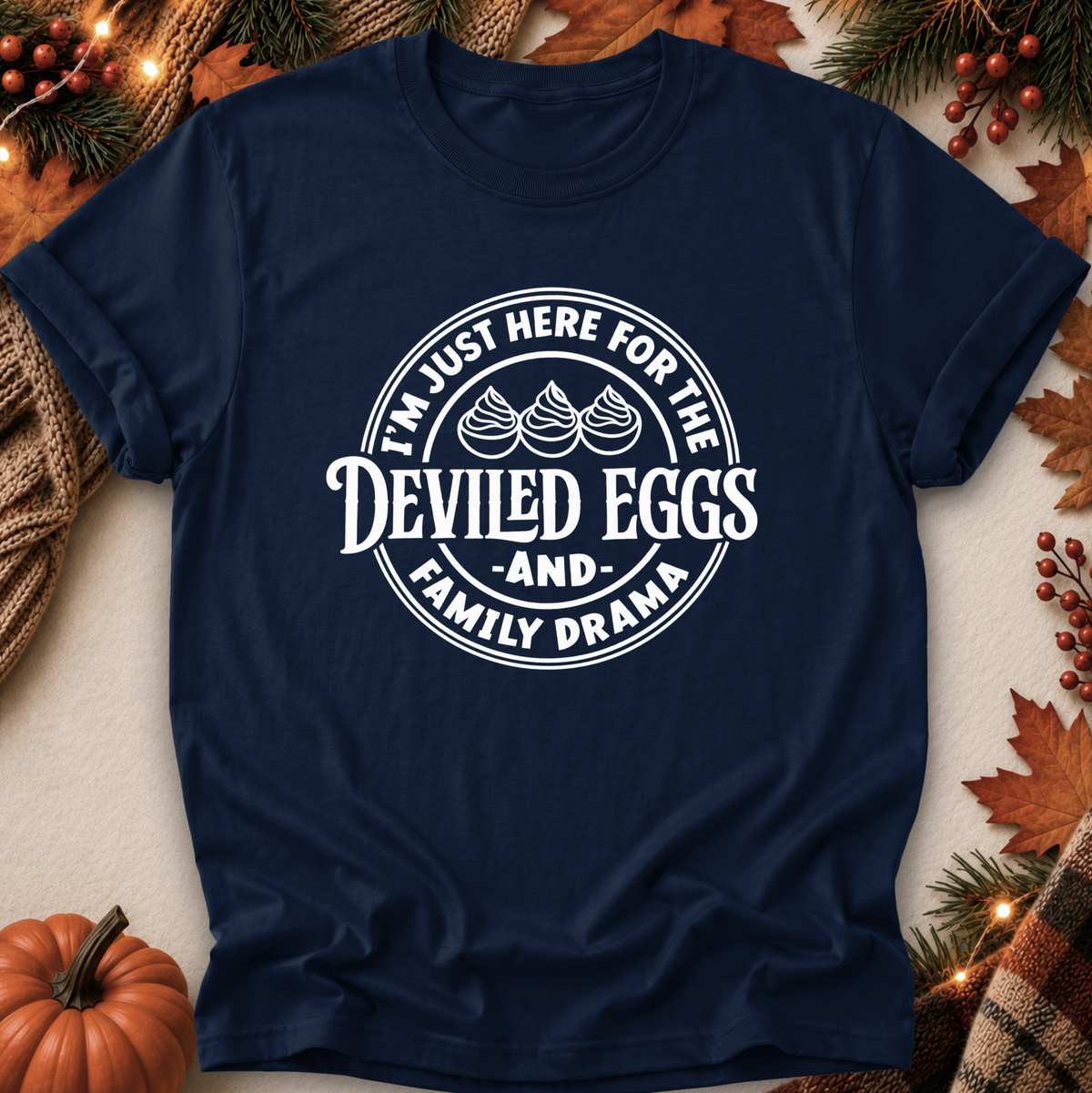 A navy blue short-sleeve t-shirt with a white circular graphic reading 'I'M JUST HERE FOR THE DEVILED EGGS AND FAMILY DRAMA' with three egg illustrations, laid flat on a neutral background with autumn props like a pumpkin, pine branches, leaves, and string lights.