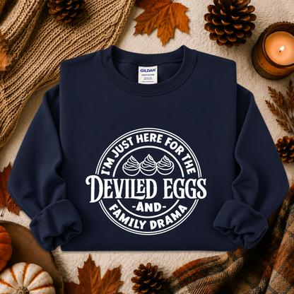 A navy blue crewneck sweatshirt with a white graphic print reading 'I'M JUST HERE FOR THE DEVILED EGGS AND FAMILY DRAMA' with three devil eggs, styled in an autumn flat-lay with a candle, pinecones, leaves, and a knit blanket.