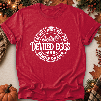 A red short-sleeve crewneck t-shirt with a white circular graphic reading 'I'M JUST HERE FOR THE DEVILED EGGS AND FAMILY DRAMA' with three egg illustrations, styled in a flat lay with autumn props like a pumpkin, pine branches, and leaves.