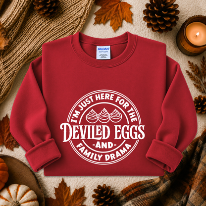 A red crewneck sweatshirt with a white graphic print reading 'I'M JUST HERE FOR THE DEVILED EGGS AND FAMILY DRAMA' with three piped eggs, styled in a flat lay with autumn props including pinecones, leaves, a candle, and a knit blanket. The Gildan brand tag is visible inside the collar.