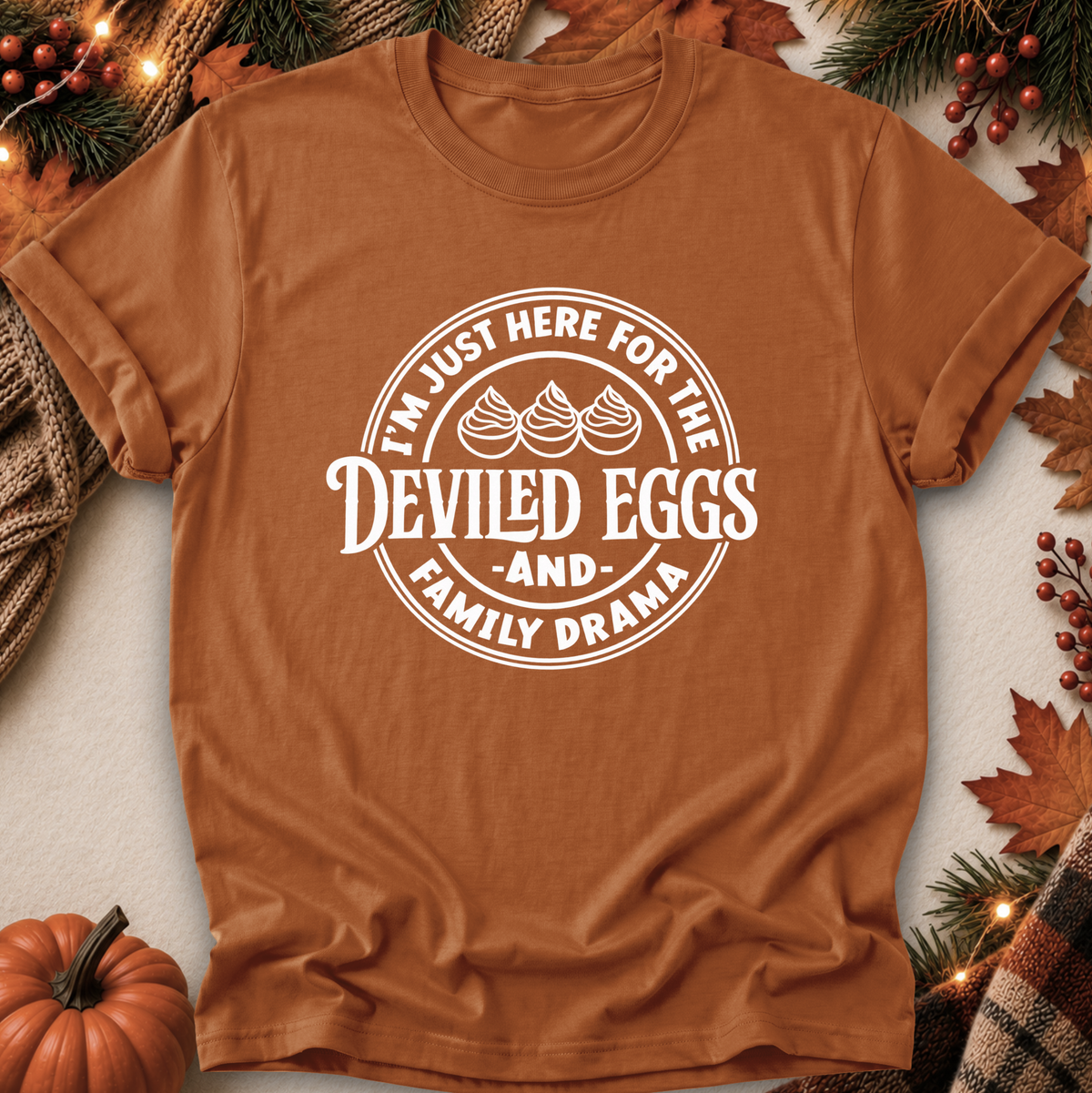 A rust-brown short-sleeve t-shirt with a white circular graphic reading 'I'M JUST HERE FOR THE DEVILED EGGS AND FAMILY DRAMA' with three egg illustrations, laid flat on a light surface with autumn decorations including a pumpkin, pine branches, leaves, and berries around it.