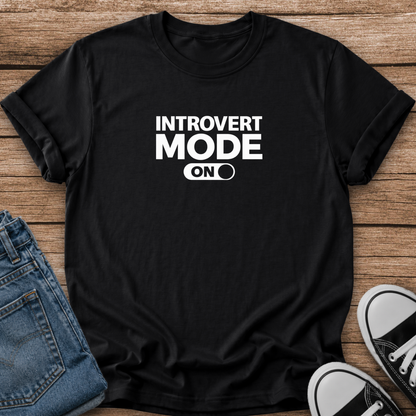 A black short-sleeve graphic T-shirt laid flat on a wooden surface, styled with folded blue jeans and black-and-white sneakers. The shirt has bold white text reading 'INTROVERT MODE ON' with a toggle switch graphic.