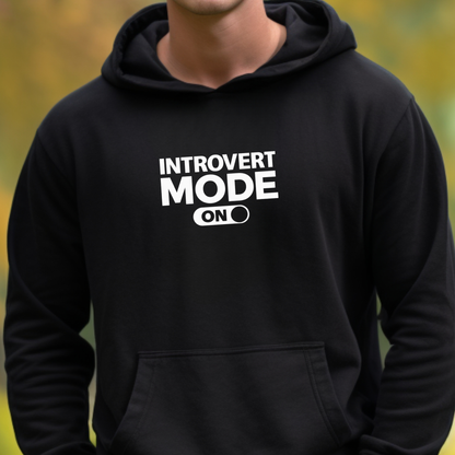 A person wearing a black pullover hoodie with white text on the chest that reads 'INTROVERT MODE ON'. The background is a soft, blurred outdoor scene with green and yellow tones.