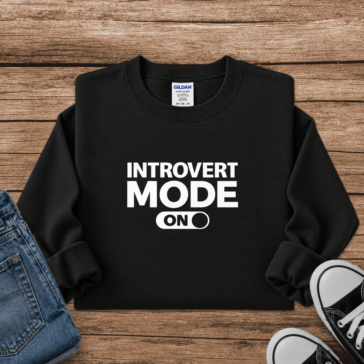 A black crewneck sweatshirt with the text 'INTROVERT MODE ON' printed in bold white letters, styled in a flat lay on a wooden background with blue jeans and black sneakers partially visible.