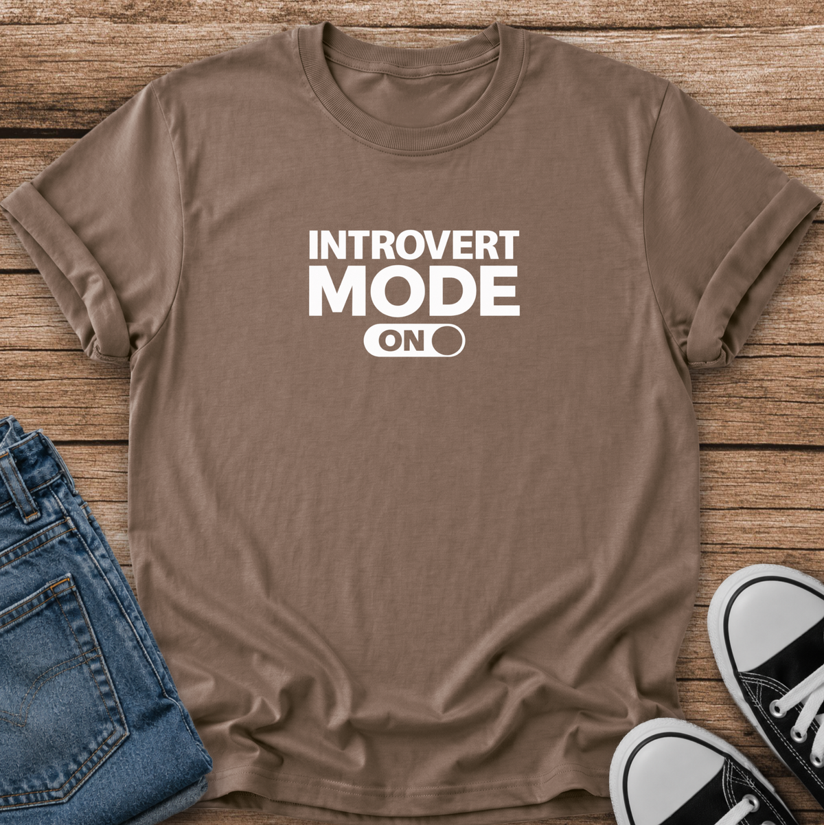 A brown short-sleeve t-shirt with white text reading 'INTROVERT MODE ON' laid flat on a rustic wood background, styled with blue jeans and black-and-white sneakers.