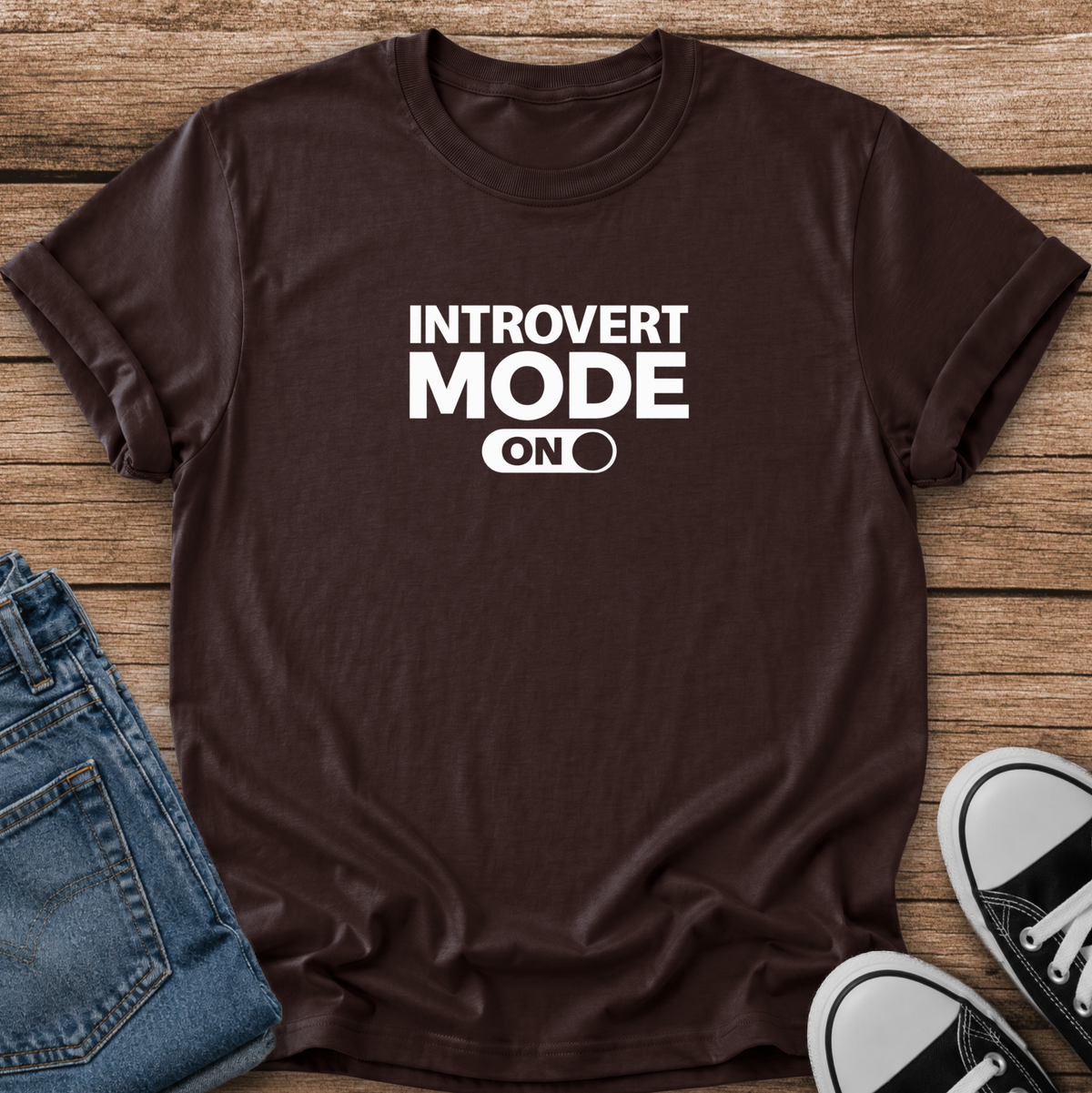 A black short-sleeve graphic T-shirt laid flat on a wooden surface, styled with folded blue jeans and black-and-white sneakers. The shirt has bold white text reading 'INTROVERT MODE ON' with a toggle switch graphic.
