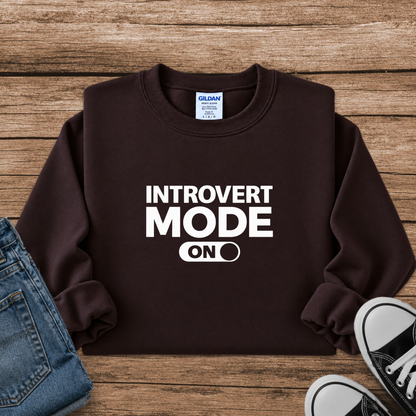 A dark brown crewneck sweatshirt with white text reading 'INTROVERT MODE ON' laid flat on a rustic wooden background, styled with a pair of blue jeans and black sneakers.