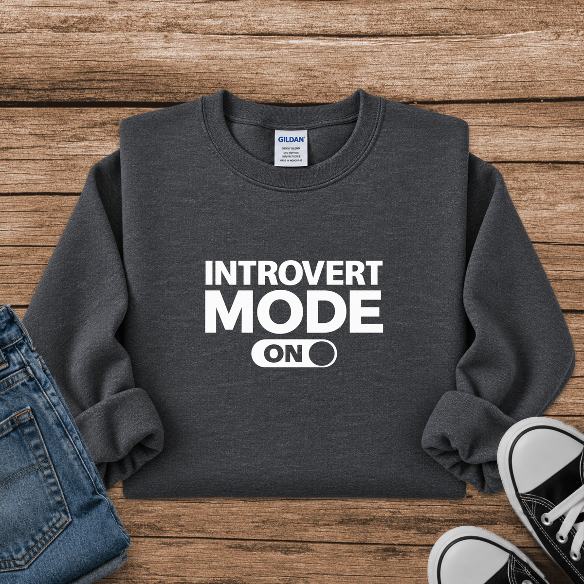 A dark gray crewneck sweatshirt with 'INTROVERT MODE ON' printed in white on the front, styled in a flat lay on a wooden background with blue jeans and black sneakers partially visible.