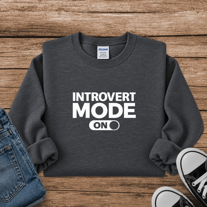 A dark gray crewneck sweatshirt with 'INTROVERT MODE ON' printed in white on the front, styled in a flat lay on a wooden background with blue jeans and black sneakers partially visible.