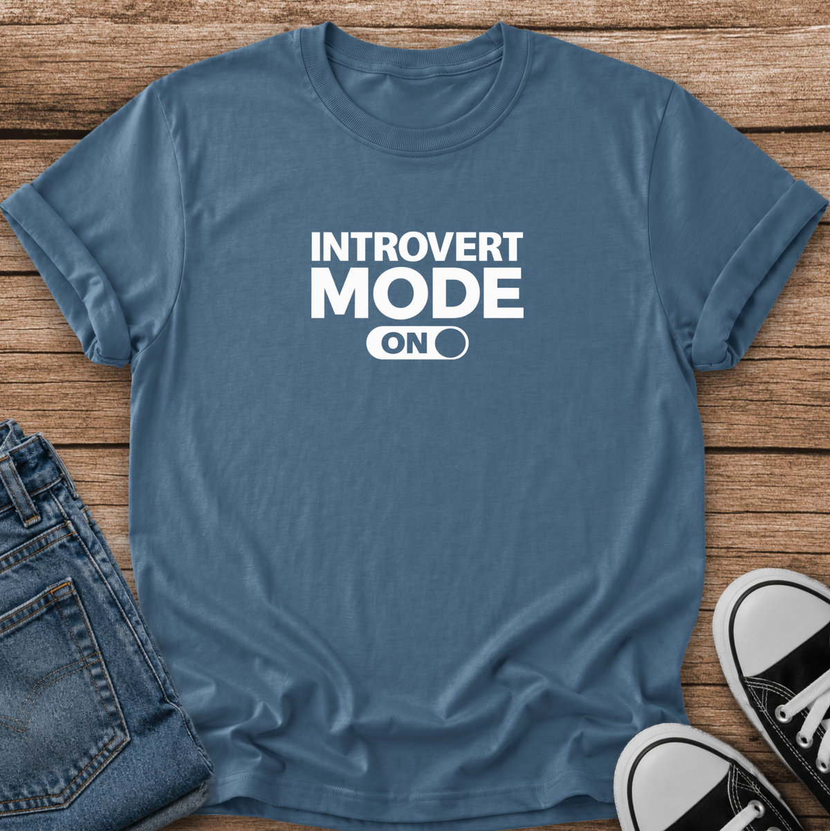 A slate blue graphic t-shirt with the text 'INTROVERT MODE ON' in white, styled in a flat lay on a wooden background with folded blue jeans and black-and-white sneakers.