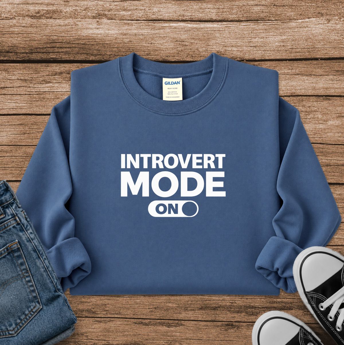 A folded blue crewneck sweatshirt with white text reading 'INTROVERT MODE ON' laid flat on a rustic wood background, styled with folded blue jeans on the left and black-and-white sneakers on the right. The neck label shows Gildan.