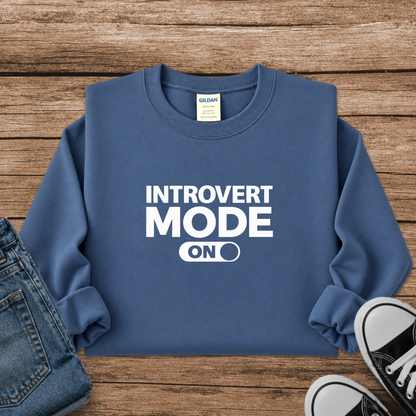 A folded blue crewneck sweatshirt with white text reading 'INTROVERT MODE ON' laid flat on a rustic wood background, styled with folded blue jeans on the left and black-and-white sneakers on the right. The neck label shows Gildan.