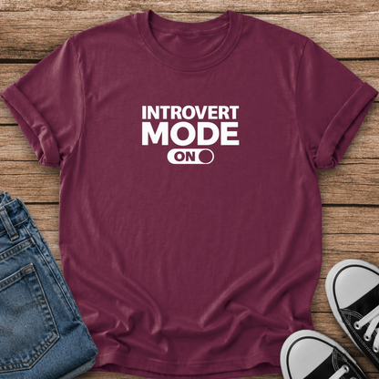 A maroon short-sleeve graphic T-shirt laid flat on a wooden surface, styled with folded blue jeans and black-and-white sneakers. The shirt has bold white text reading 'INTROVERT MODE ON' with a toggle switch graphic.