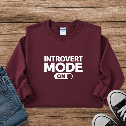A maroon crewneck sweatshirt with white text reading 'INTROVERT MODE ON' is laid flat on a rustic wood background, styled with a pair of blue jeans and black sneakers.
