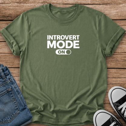 An olive green short-sleeve t-shirt with white text reading 'INTROVERT MODE ON' laid flat on a rustic wooden background, styled with folded blue jeans and black-and-white sneakers.