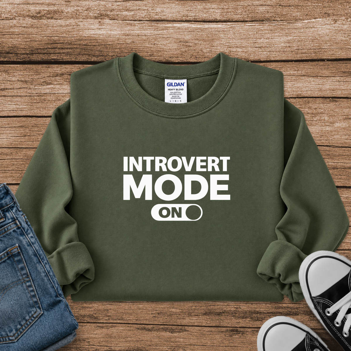 An olive green crewneck sweatshirt with 'INTROVERT MODE ON' printed in bold white text on the front, styled in a flat lay on a wooden background with blue jeans and black sneakers.