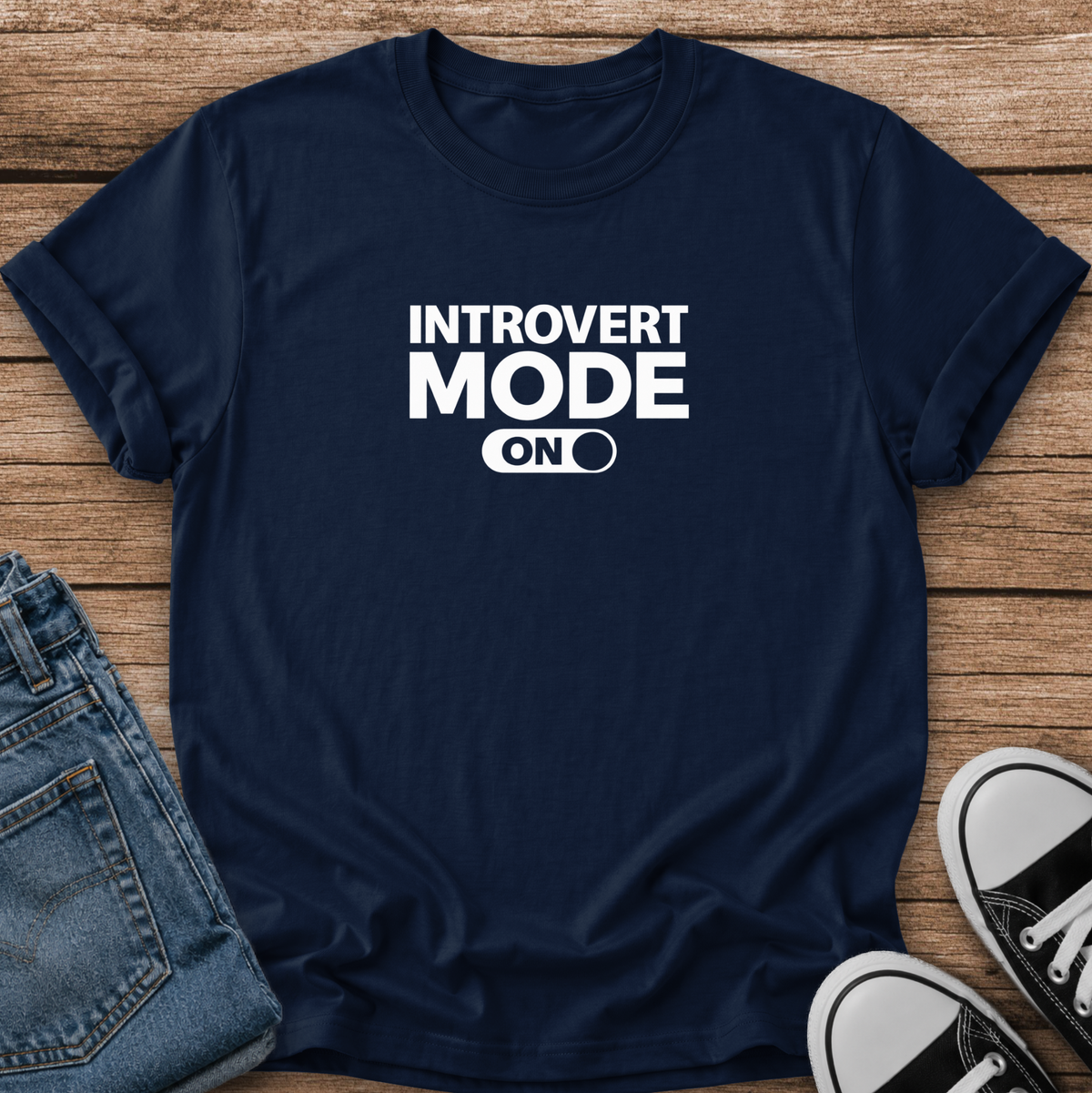 A navy blue short-sleeve t-shirt with white text reading 'INTROVERT MODE ON' laid flat on a wooden surface, styled with folded blue jeans and black-and-white sneakers.