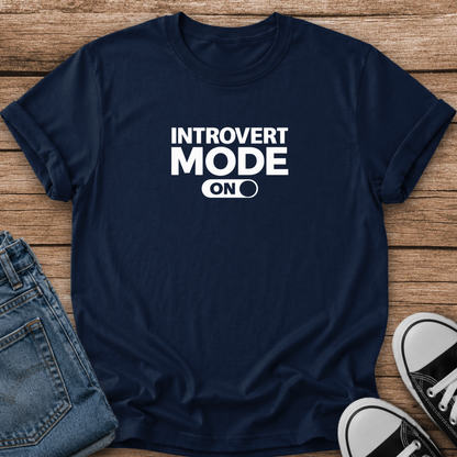 A navy blue short-sleeve t-shirt with white text reading 'INTROVERT MODE ON' laid flat on a wooden surface, styled with folded blue jeans and black-and-white sneakers.