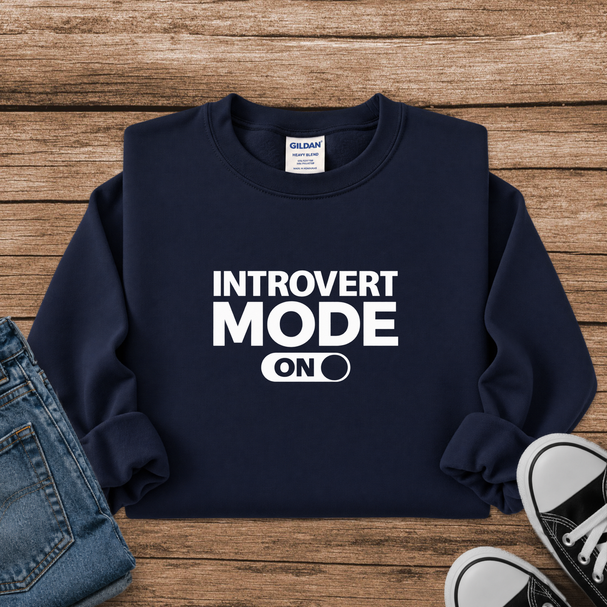A navy blue crewneck sweatshirt with 'INTROVERT MODE ON' printed in bold white text on the front, styled in a flat lay on a wooden surface with jeans and black-and-white sneakers partially visible.
