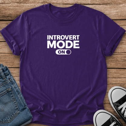 A purple short-sleeve graphic T-shirt laid flat on a wood surface, with folded blue jeans and black-and-white sneakers partly visible around it. The shirt has bold white text reading 'INTROVERT MODE ON' across the chest.