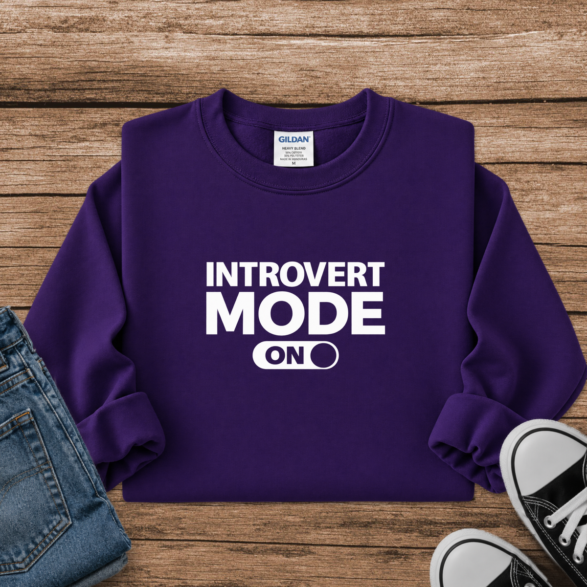 A purple long-sleeve sweatshirt with white text reading 'INTROVERT MODE ON' laid flat on a wooden surface, styled with folded blue jeans and black-and-white sneakers.