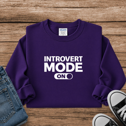 A purple long-sleeve sweatshirt with white text reading 'INTROVERT MODE ON' laid flat on a wooden surface, styled with folded blue jeans and black-and-white sneakers.