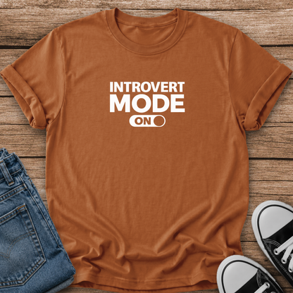 A rust-orange crewneck t-shirt with white text reading 'INTROVERT MODE ON' laid flat on a rustic wood background, styled with blue jeans and black-and-white sneakers.