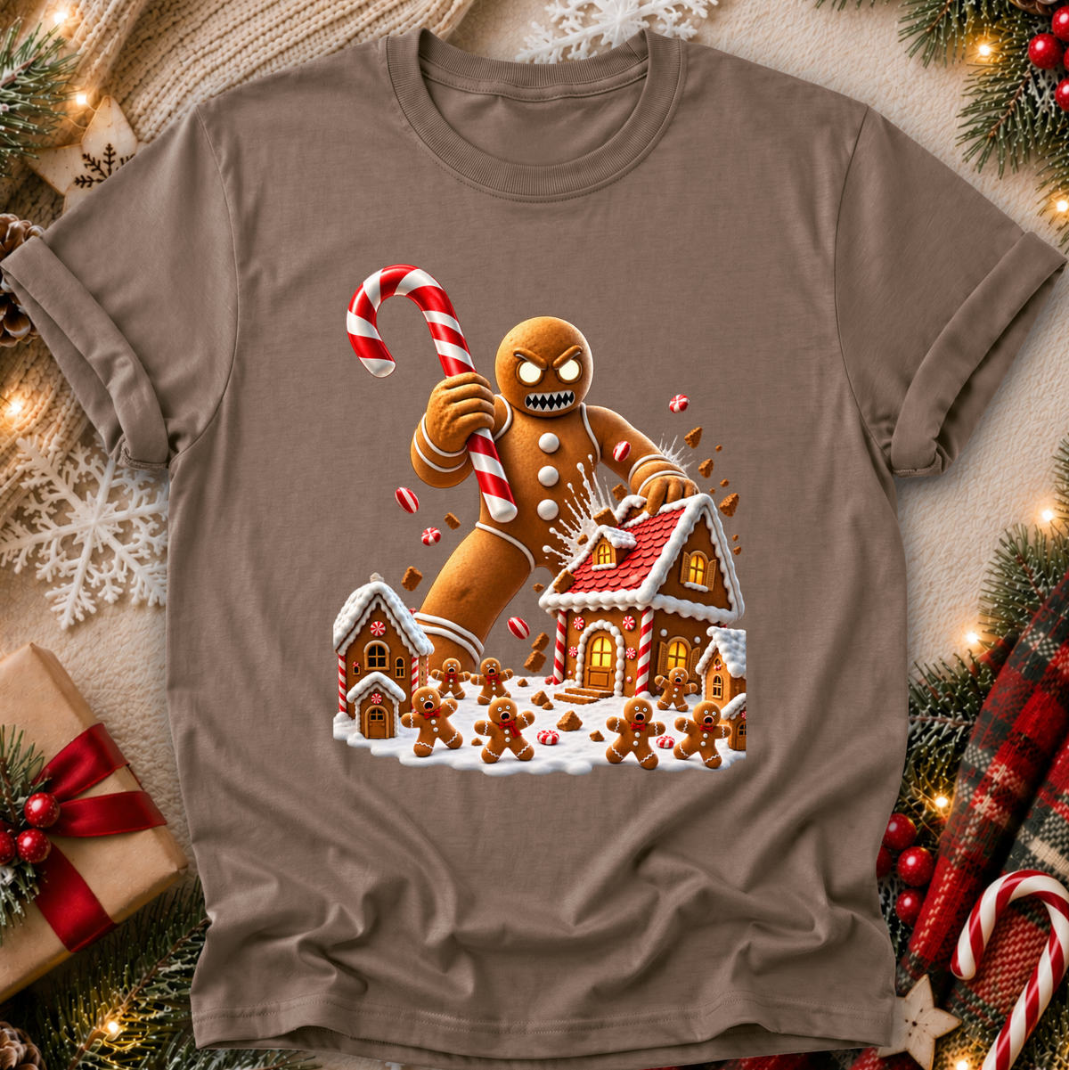 A taupe brown short-sleeve crewneck t-shirt featuring a festive Christmas gingerbread graphic, laid flat on a cozy holiday-themed background with pine branches, lights, and a gift box.