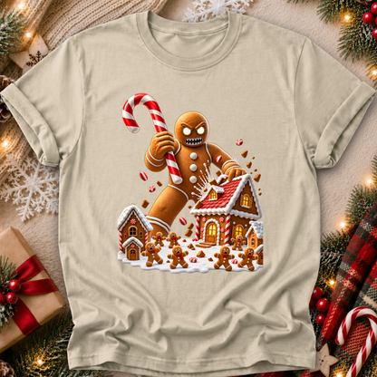 A beige short-sleeve crewneck t-shirt featuring a festive Christmas graphic of a gingerbread man smashing a gingerbread house while holding a candy cane, surrounded by holiday decorations like pine branches, lights, and a wrapped gift.