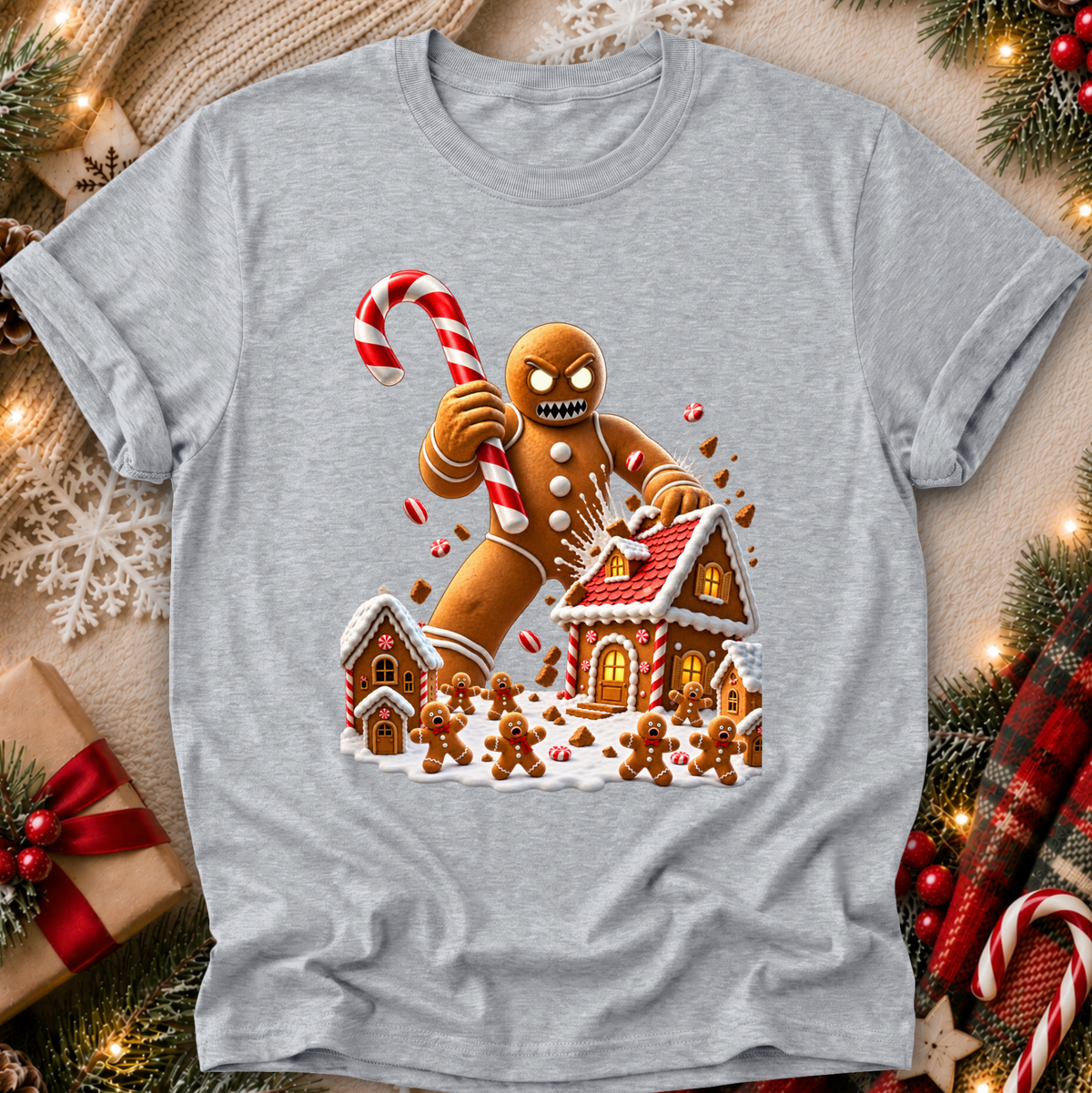 A heather gray short-sleeve t-shirt with a festive Christmas graphic of a gingerbread man holding a candy cane in front of gingerbread houses. The t-shirt is displayed in a flat-lay setting surrounded by holiday decorations including pine branches, a wrapped gift, and a plaid scarf.