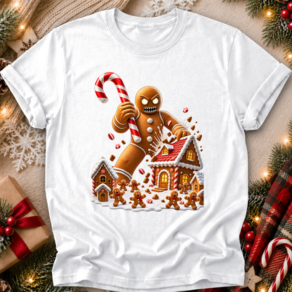 A white short-sleeve crewneck t-shirt featuring a festive Christmas evil gingerbread graphic, laid flat on a cozy holiday-themed background with pine branches, lights, and a gift box.