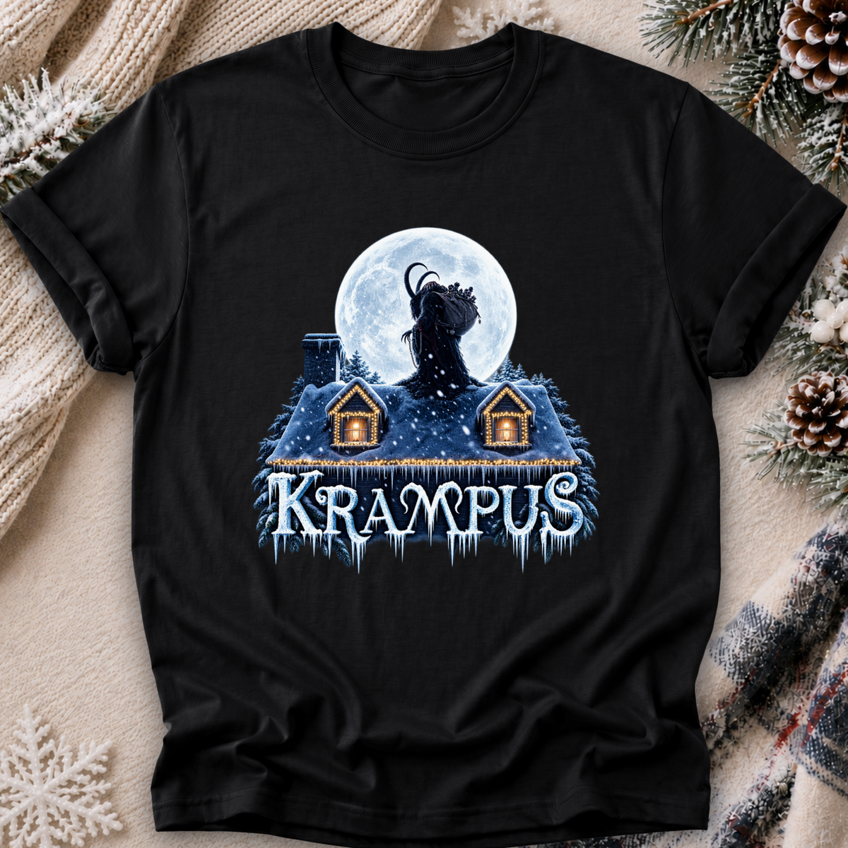 A black short-sleeve graphic T-shirt laid flat on a cozy beige knit blanket with pinecones and evergreen branches around it. The shirt features a winter-themed graphic of a goat-like figure in front of a full moon above snow-covered roofs, with the word 'Krampus' in icy blue lettering.