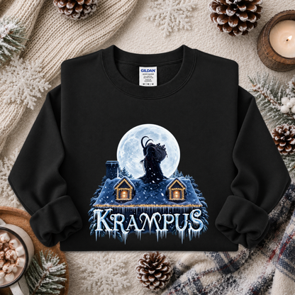 A black long-sleeve sweatshirt laid flat on a cozy holiday-themed background with pinecones, a candle, and knit fabric. The sweatshirt features a graphic of a dark goat-like figure in front of a full moon over snow-covered roofs, with the word 'Krapus' in icy blue lettering below.