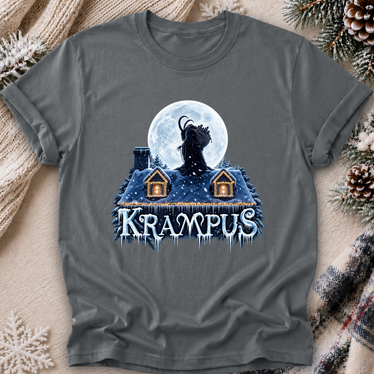 A dark gray graphic t-shirt featuring a spooky winter illustration of a goat-like figure in front of a full moon over a snow-covered house, with the word 'Krampus' in icy blue letters. The shirt is laid flat on a cozy background with a knitted blanket, pinecones, and evergreen branches.
