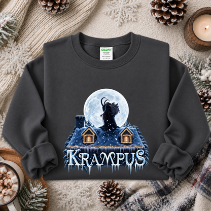 A dark charcoal gray crewneck sweatshirt featuring a festive winter graphic of a Krampus figure in front of a full moon above snow-covered houses, with the word 'KRAMPUS' in icy blue letters. The sweatshirt is styled in a cozy flat-lay arrangement with pinecones, a knitted blanket, and a candle.
