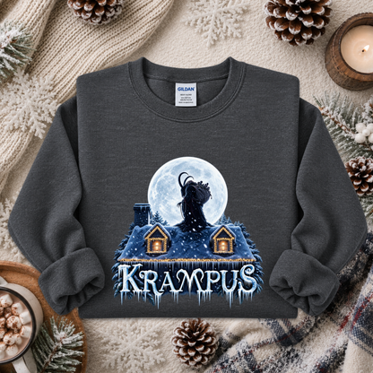 A dark gray crewneck sweatshirt featuring a winter-themed graphic of a goat silhouette against a full moon above snow-covered houses, with the word 'KRAMPUS' in icy blue letters. The sweatshirt is styled in a cozy flat-lay arrangement with a knitted beige scarf, pinecones, a lit candle, and a mug of hot cocoa.